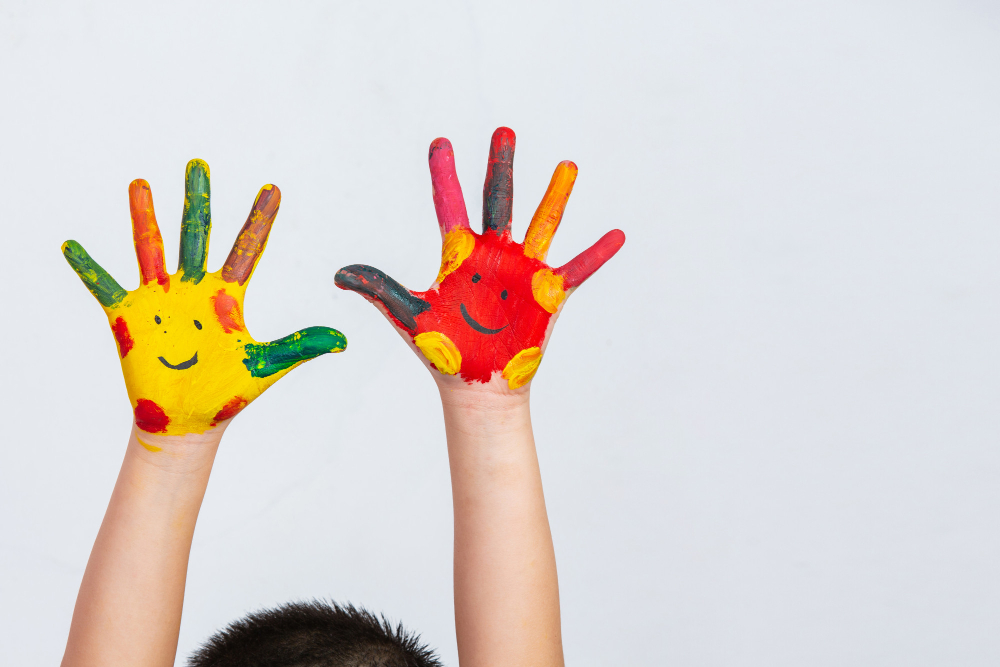 Foto. Fundo cinza claro, dois antebraços de uma criança com as mãos bem abertas e as palmas viradas para frente. A pele dela é branca e as mãos estão pintadas de tinta colorida (amarelo, vermelho, laranja, rosa, verde, marrom), cada uma com dois olhos e um sorriso desenhados com tinta preta. É possível ver também o topo da cabeça da criança, ela tem o cabelo curto e preto.