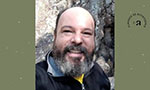 Composição em formato retangular. No centro, uma foto quadrada de um homem branco e adulto, ele tem barba parcialmente grisalha, está sorrindo e usa uma jaqueta preta; no fundo uma parede de pedra parte bege claro e parte cinza. Nos lados da foto, dois retângulos na vertical em tom verde musgo com pequenos detalhes brancos. No retângulo direito, um logo na cor preta (a letra “e” bem pequena, um traço, a letra “a” com aproximadamente o triplo do tamanho da letra “e”, em volta formando um arco o texto “Almanack no Bicentenário”).