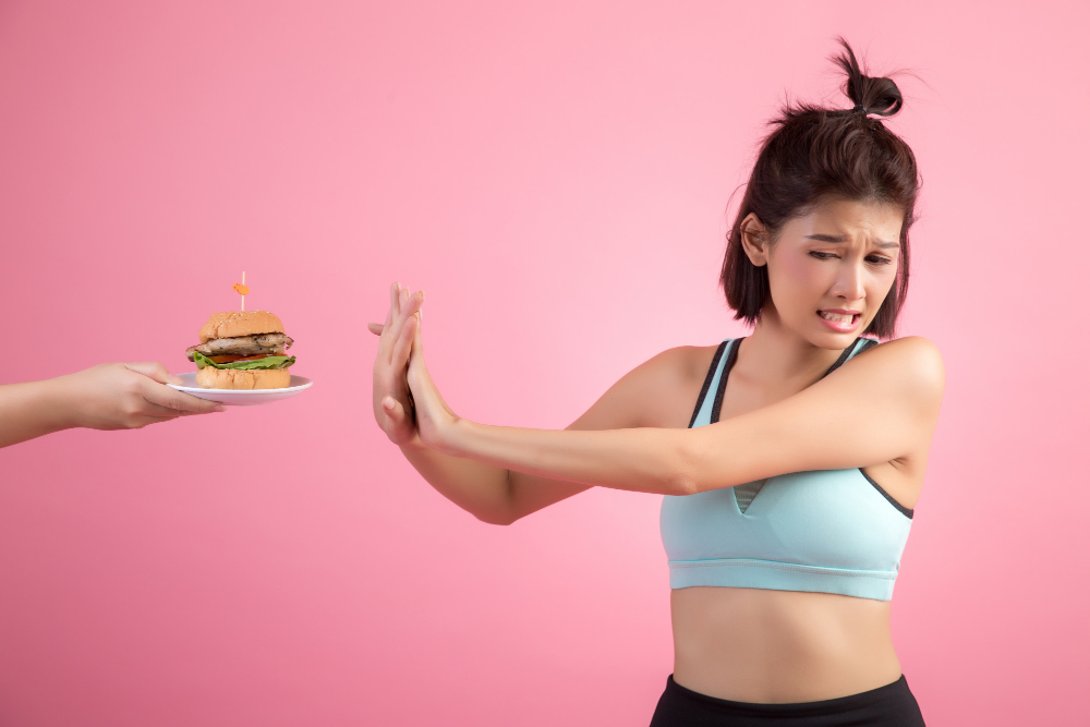Foto retangular horizontal. Uma mulher adulta recusa um hambúrguer. Ela faz uma careta, estende os braços na direção do hambúrguer e sobrepõe as duas mãos. Ela usa roupa de ginástica. Fundo rosa difuso.