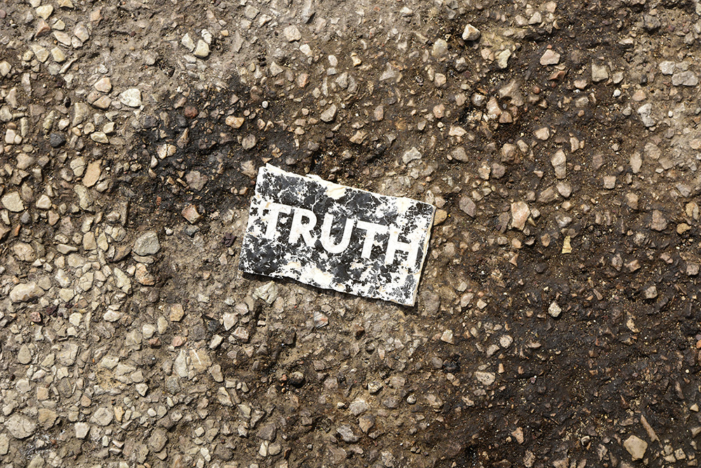 Foto retangular horizontal. Papel retangular na horizontal com a palavra TRUTH (verdade, em inglês) muito desgastado no chão. O papel é preto ou cinza escuro e as letras brancas. No chão pedregulhos e terra.