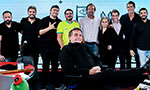 Foto: Bolsonaro está sorrindo e sentado em uma cadeira com os pés sobre a mesa. Atrás, equipe da Jovem Pan lado a lado. De nove pessoas, apenas dois não estão sorrindo. O lugar parece ser um estúdio, com iluminação forte, cenário e microfones.