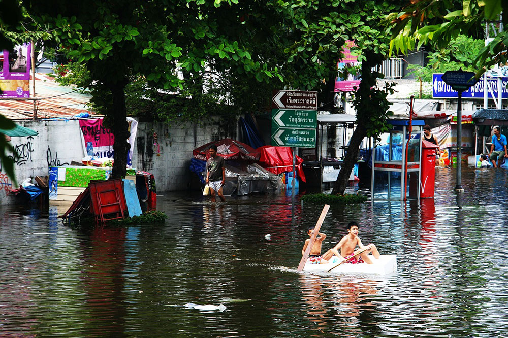 Foto de uma rua completamente alagada. Dois meninos dentro de uma espécie de caixa ou barco e remando com tábuas. No fundo: um muro, árvores, pessoas, placas de sinalização e barracas de comida cercadas por água.