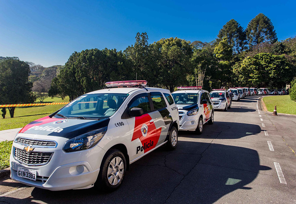 Foto: várias viaturas estatacionadas em fila na rua. No fundo, gramado, muitas árvores e céu claro.