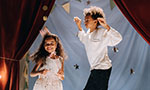 Foto: na esquerda uma menina sorrindo que parece estar dançando, na direita um menino com expressão neutra e fazendo pinça com as mãos (como um caranguejo). No fundo, um painel de tecido com estrelas, um fio com duas lâmpadas acesas e penduradas, barbantes com bandeirinhas (algumas no formato de triangulo e outras de círculo) e duas cortinas vermelhas com uma amarração na parte média inferior.