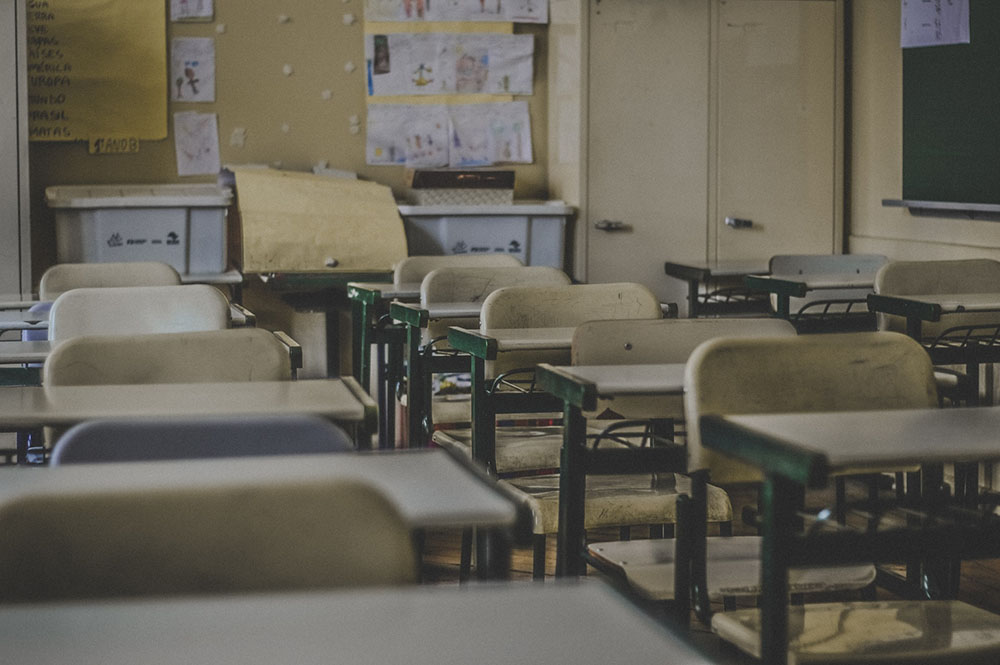 Foto de uma sala de aula com várias cadeiras enfileiradas. No fundo, armários e desenhos colados na parede. O tom da foto está meio escurecido, dando a impressão de ser um lugar abandonado.