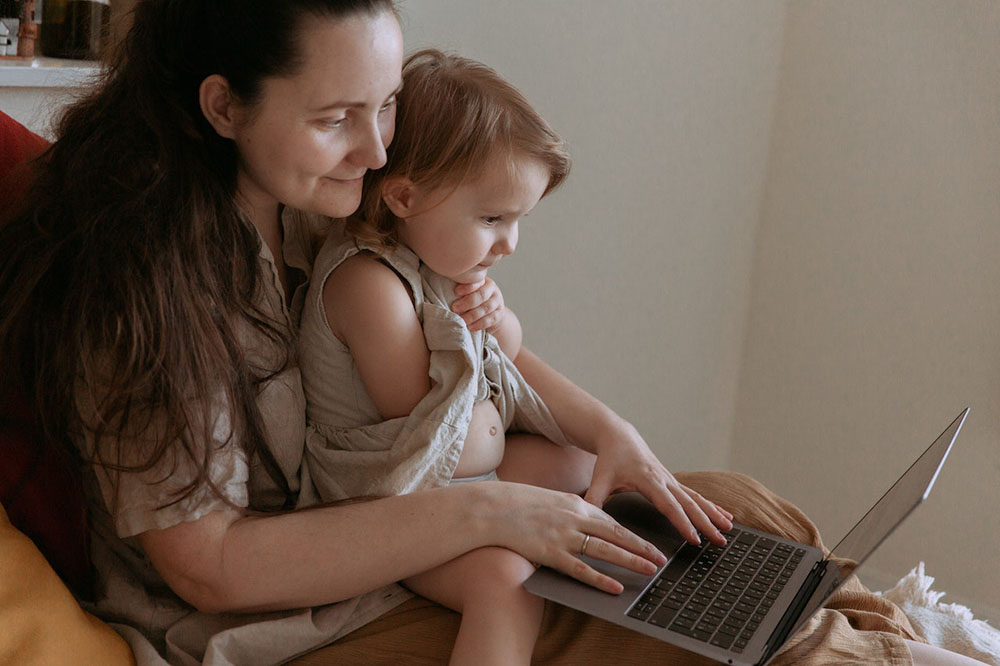 Foto de uma mulher sentada com uma criança pequena no colo e um notebook sobre as pernas.