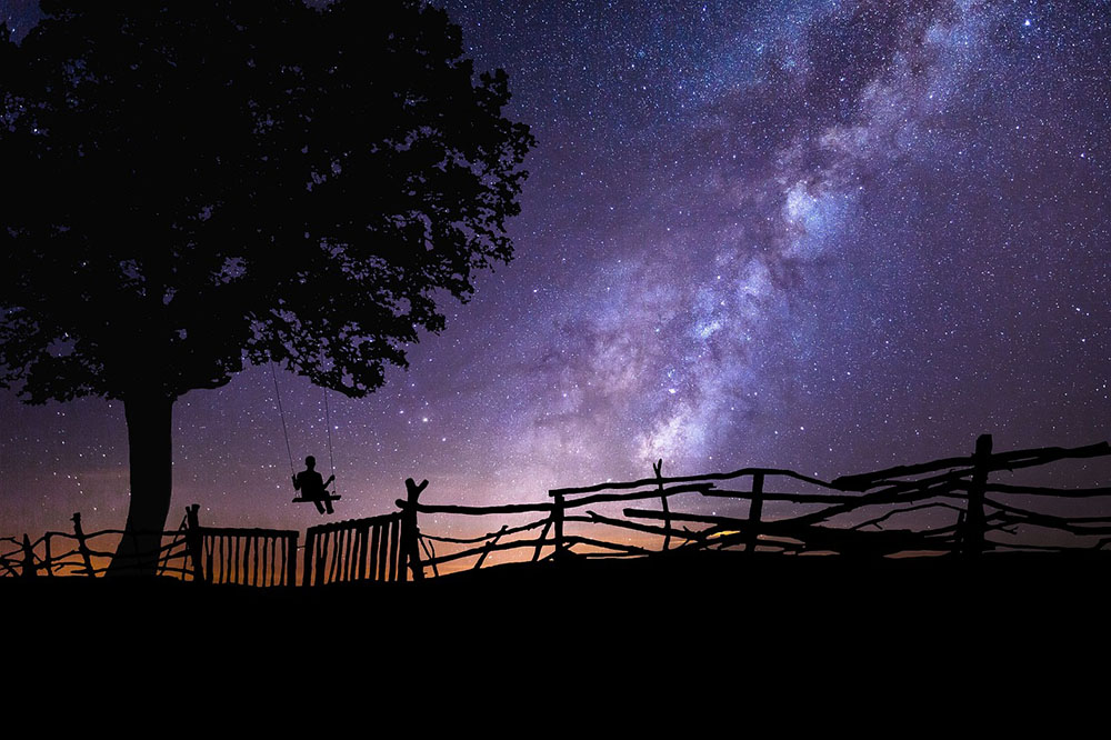 Composição. Céu estrelado em tons roxo e laranja. Sombra de uma pessoa sentada em um balanço, uma árvore e cercas. Composição. Céu estrelado em tons roxo e laranja. Sombra de uma pessoa sentada em um balanço, uma árvore e cercas.