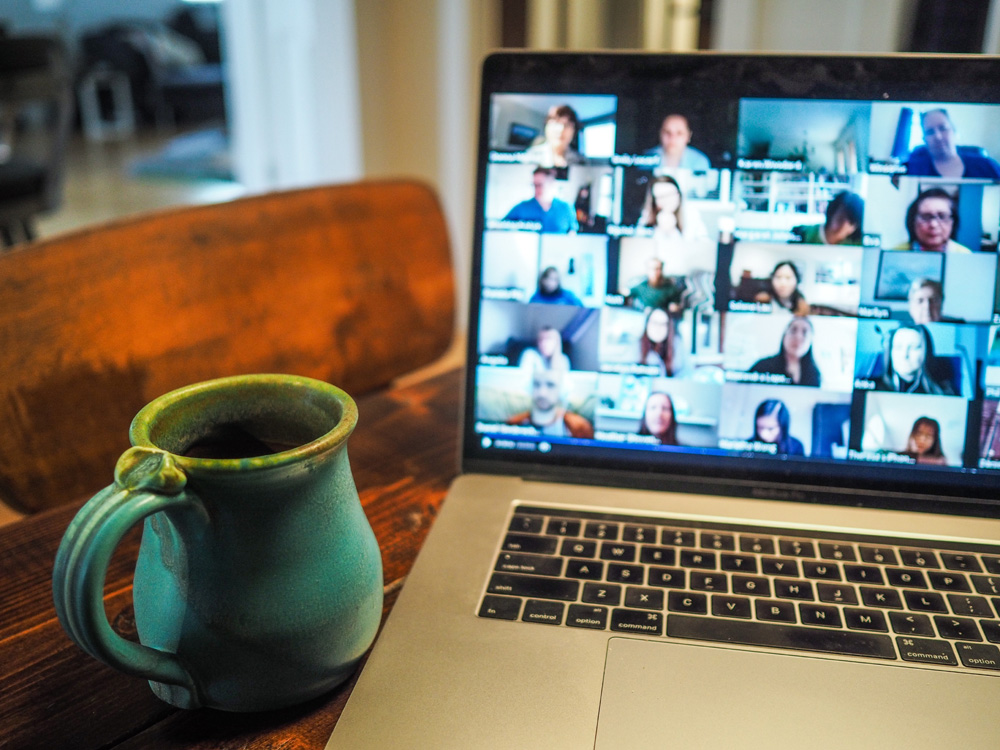 Notebook sobre uma mesa de madeira. A tela apresenta uma videoconferência. Ao lado do aparelho está uma caneca de porcelana azul, com borda verde. O fundo da imagem está desfocado. Notebook sobre uma mesa de madeira. A tela apresenta uma videoconferência. Ao lado do aparelho está uma caneca de porcelana azul, com borda verde. O fundo da imagem está desfocado.