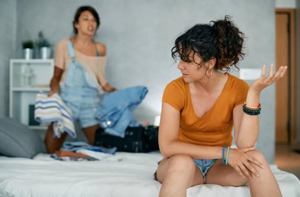 Duas mulheres estão discutindo. A que está mais à frente, sentada em uma cama, está olhando de perfil para a outra, que está em pé, mais ao fundo. Ela segura algumas roupas, com expressão de raiva, enquanto encara a primeira mulher. Duas mulheres estão discutindo. A que está mais à frente, sentada em uma cama, está olhando de perfil para a outra, que está em pé, mais ao fundo. Ela segura algumas roupas, com expressão de raiva, enquanto encara a primeira mulher.