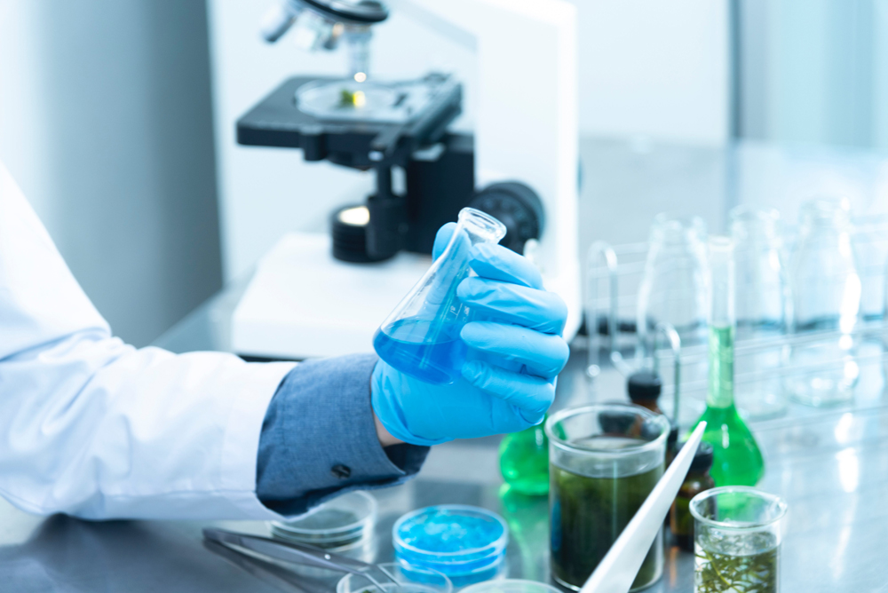 Fotografia de uma mão segurando um frasco em um laboratório, que pode ser visto ao fundo. Fotografia de uma mão segurando um frasco em um laboratório, que pode ser visto ao fundo.