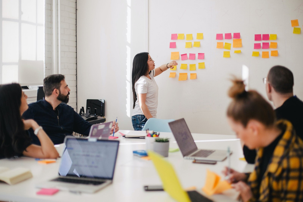 Grupo de cinco pessoas (três mulheres e dois homens) em reunião em uma sala. Quatro estão sentadas em uma mesa com notebooks e agendas, enquanto uma mulher em pé à frente da sala observa e aponta para post-its fixados na parede. Grupo de cinco pessoas (três mulheres e dois homens) em reunião em uma sala. Quatro estão sentadas em uma mesa com notebooks e agendas, enquanto uma mulher em pé à frente da sala observa e aponta para post-its fixados na parede.