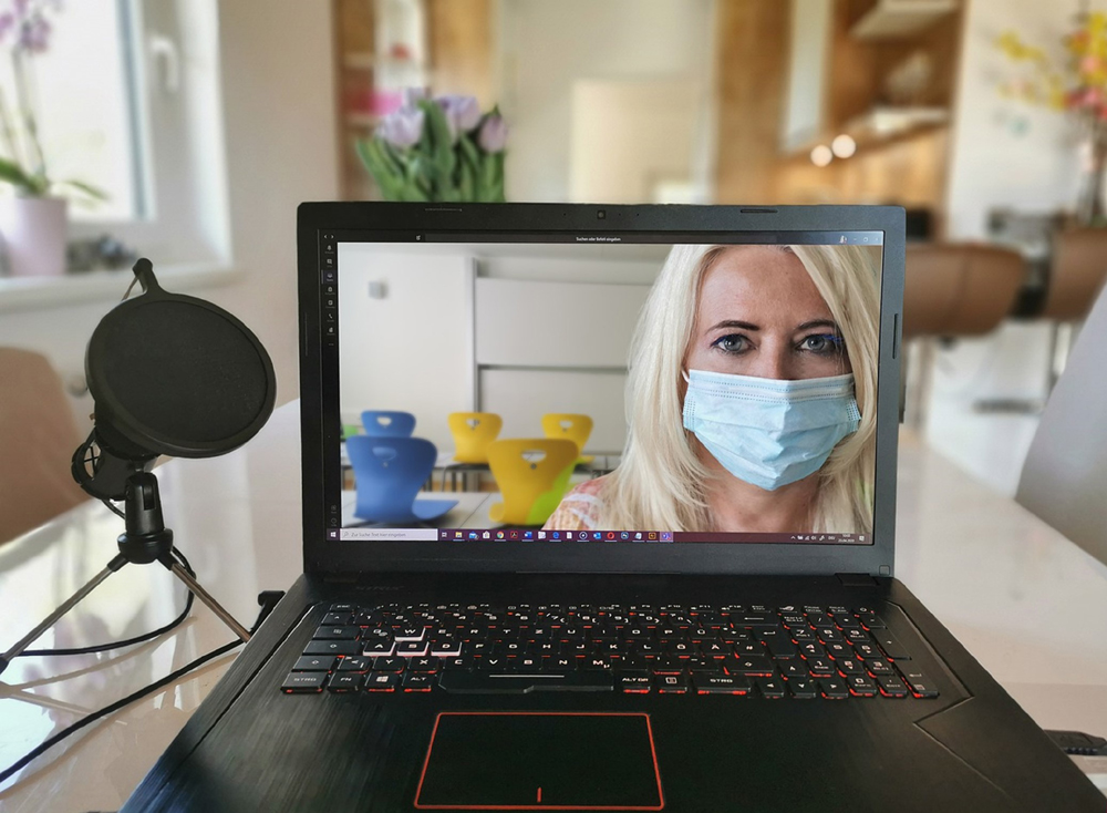 Fotografia de um notebook ligado, exibindo uma mulher loira com máscara na tela. O dispositivo repousa sobre uma mesa branca espelhada, ao lado de um microfone de mesa. Ao fundo, há uma sala ampla com alguns móveis, que está desfocada. Fotografia de um notebook ligado, exibindo uma mulher loira com máscara na tela. O dispositivo repousa sobre uma mesa branca espelhada, ao lado de um microfone de mesa. Ao fundo, há uma sala ampla com alguns móveis, que está desfocada.