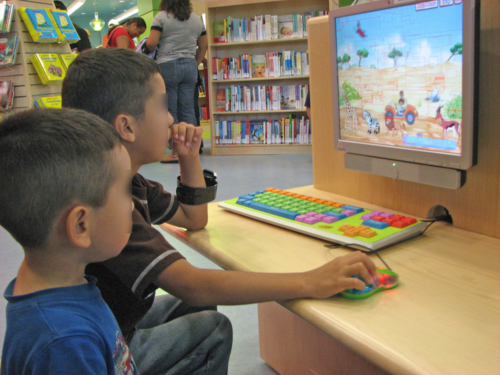 Dois meninos em uma biblioteca jogando em um computador. Ao fundo, há prateleiras com livros, ao lado de duas mulheres adultas, que estão em pé. Dois meninos em uma biblioteca jogando em um computador. Ao fundo, há prateleiras com livros, ao lado de duas mulheres adultas, que estão em pé.