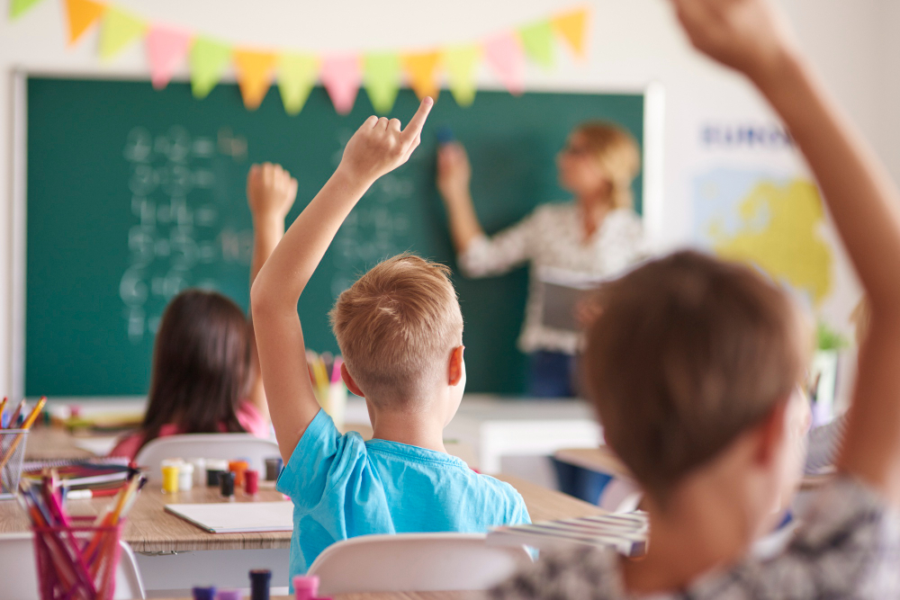 Sala de aula com três crianças de costas, erguendo a mão, como se procurassem responder a alguma pergunta da professora, que está à frente da sala, apontando para a lousa. Sala de aula com três crianças de costas, erguendo a mão, como se procurassem responder a alguma pergunta da professora, que está à frente da sala, apontando para a lousa.