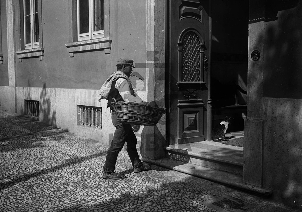 Fotografia em preto e branco de um padeiro vendendo pão a domicílio. O homem usa uma boina, calça escura e um bolero preto sobre uma camisa branca de manga longa. Ele está posicionado em frente à porta de uma residência, segurando um cesto grande em seu braço direito. Fotografia em preto e branco de um padeiro vendendo pão a domicílio. O homem usa uma boina, calça escura e um bolero preto sobre uma camisa branca de manga longa. Ele está posicionado em frente à porta de uma residência, segurando um cesto grande em seu braço direito.