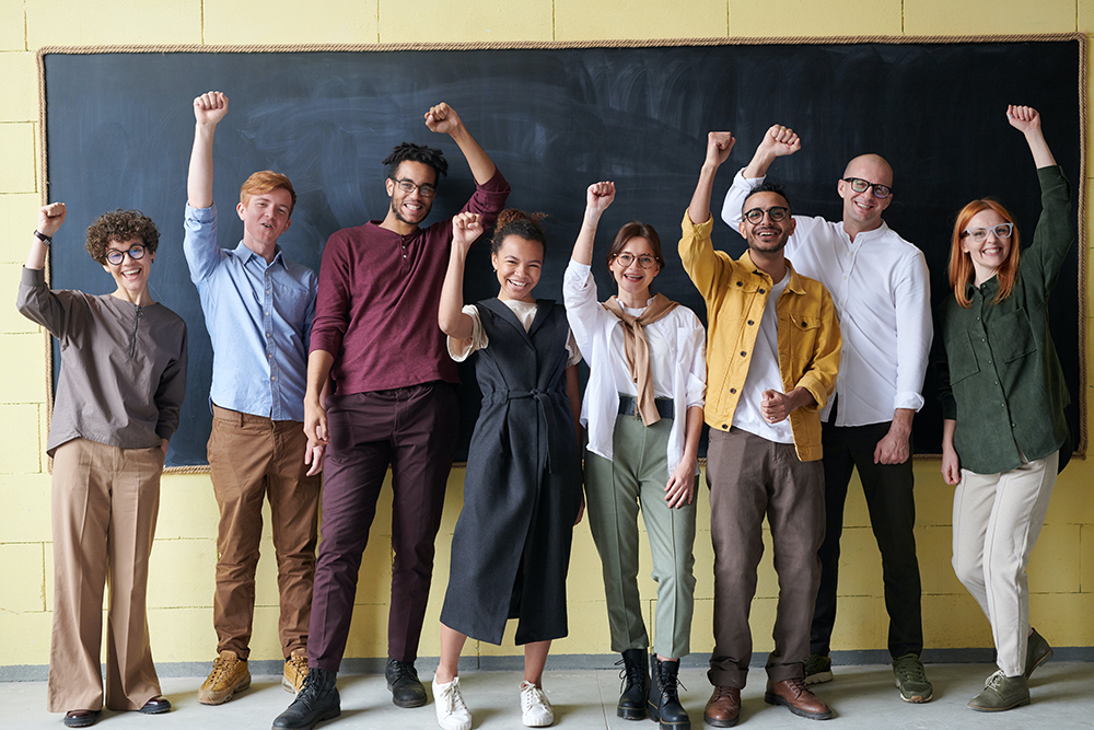 Um grupo diversificado de oito pessoas sorrindo e levantando os braços em comemoração, em frente a um quadro negro. Um grupo diversificado de oito pessoas sorrindo e levantando os braços em comemoração, em frente a um quadro negro.