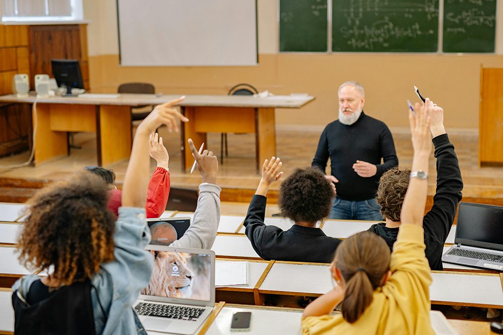 Professor de meia-idade ensinando em uma sala de aula ampla, onde alunos estão sentados em suas carteiras, com notebooks abertos diante deles, e vários levantam as mãos para fazer perguntas Professor de meia-idade ensinando em uma sala de aula ampla, onde alunos estão sentados em suas carteiras, com notebooks abertos diante deles, e vários levantam as mãos para fazer perguntas
