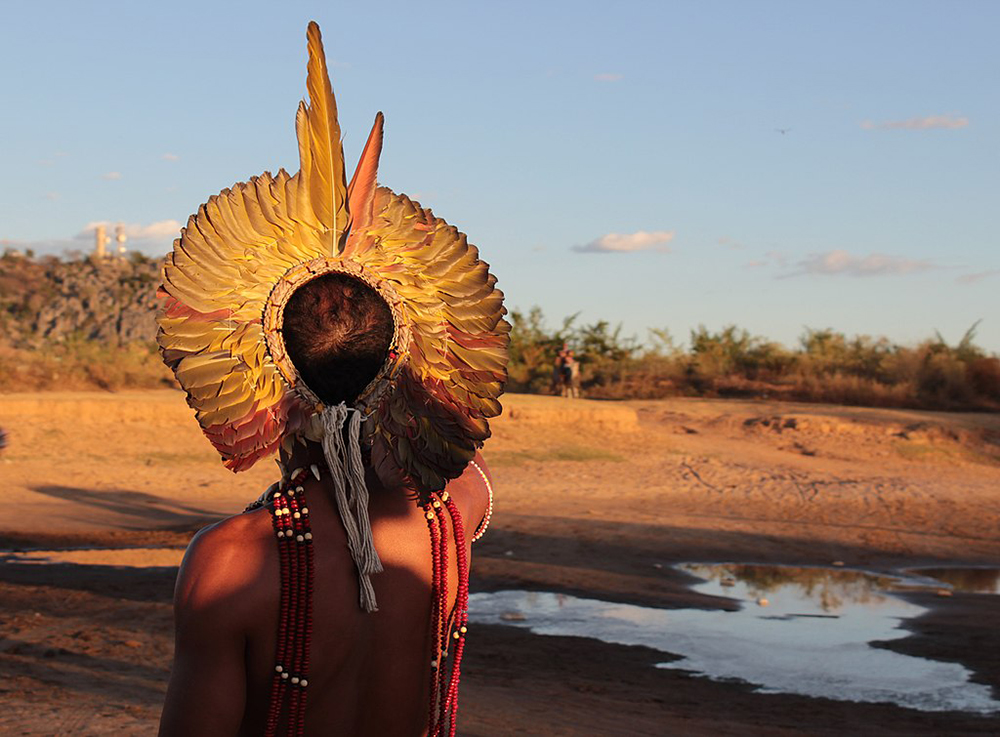 Fotografia de um homem indígena de costas, observando o horizonte. Ele usa um cocar colorido e diversos adereços tradicionais. Ao seu lado, há um pequeno lago cercado por um campo aberto com diversas vegetações. O céu acima está claro e azul. Fotografia de um homem indígena de costas, observando o horizonte. Ele usa um cocar colorido e diversos adereços tradicionais. Ao seu lado, há um pequeno lago cercado por um campo aberto com diversas vegetações. O céu acima está claro e azul.