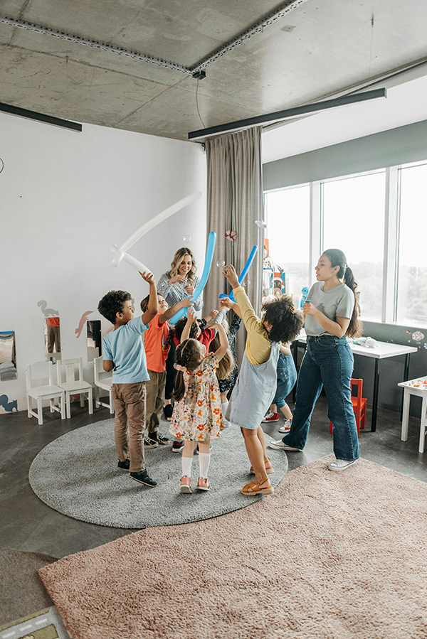 Fotografia de crianças brincando com bexigas e bolhas de sabão, supervisionadas por duas adultas, em uma sala ampla com decoração infantil e alguns tapetes dispostos no chão. Fotografia de crianças brincando com bexigas e bolhas de sabão, supervisionadas por duas adultas, em uma sala ampla com decoração infantil e alguns tapetes dispostos no chão.