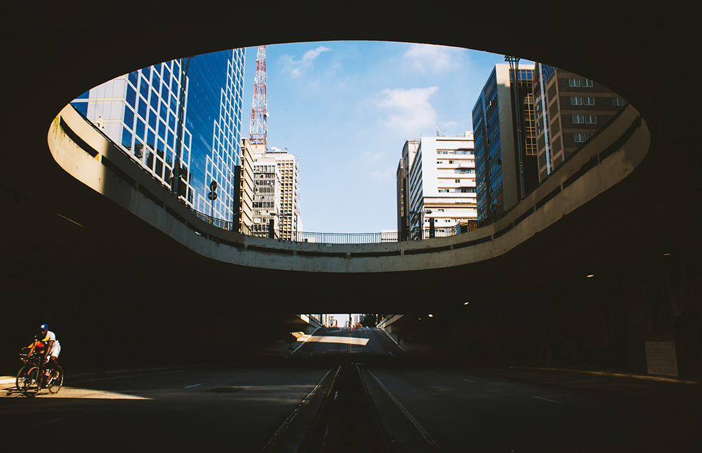 Fotografia de uma pessoa pedalando uma bicicleta dentro de um túnel. O túnel tem uma abertura circular no topo, permitindo a visão parcial dos edifícios da rua acima e do céu azul Fotografia de uma pessoa pedalando uma bicicleta dentro de um túnel. O túnel tem uma abertura circular no topo, permitindo a visão parcial dos edifícios da rua acima e do céu azul