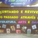 Fotografia de um painel marrom com o título "Recontando e Revivendo o Passado através da Literatura, Arte e Dança" nas cores amarela, azul, roxo e rosa. Abaixo o título, tem impressões de capas de livros infantis que remetem ao título