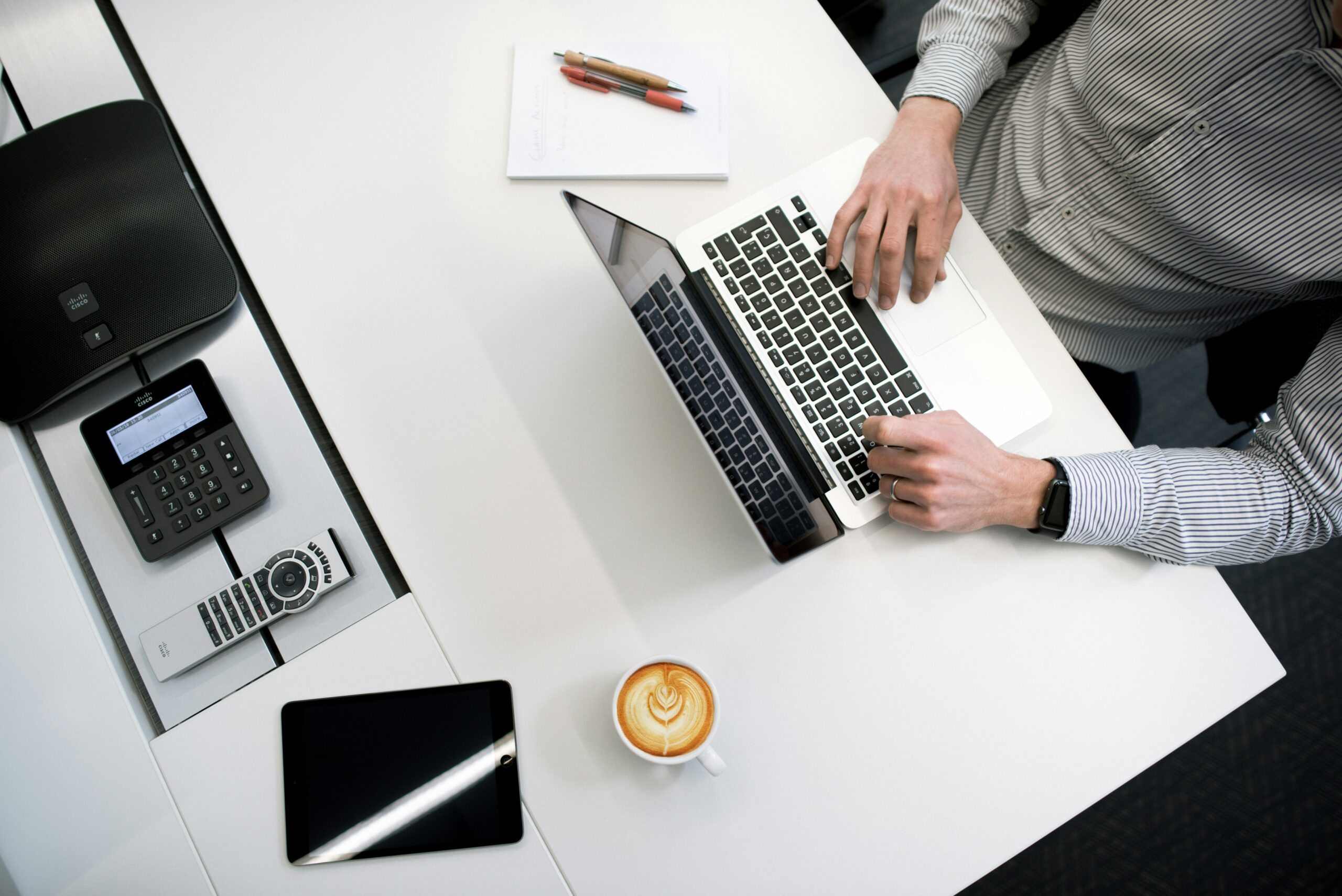 Fotografia de uma mesa branca com um notebook, ao qual um homem branco e de camisa de botões cinza está digitando, um bloco de anotações em branco com duas canetas, uma xícara com café, um tablet, uma calculadora, um controle remoto e um aparelho de som Fotografia de uma mesa branca com um notebook, ao qual um homem branco e de camisa de botões cinza está digitando, um bloco de anotações em branco com duas canetas, uma xícara com café, um tablet, uma calculadora, um controle remoto e um aparelho de som