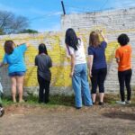 A imagem mostra cinco jovens usando roupas despojadas pintado um muro branco com tinta amarela. Usando pincéis, trabalham em equipe, dando cor ao espaço. A cena transmite colaboração e criatividade, em um ambiente urbano, iluminado pela luz do dia.
